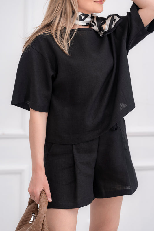 lininiai šortai moterims, trumpi, platūs, su klostėmis priekyje. Liemuo su guma ir raištuku, šortai su kišenėmis. Spalva - juoda. Modelis apsivilkusi ir derančią lininę palaidinę.