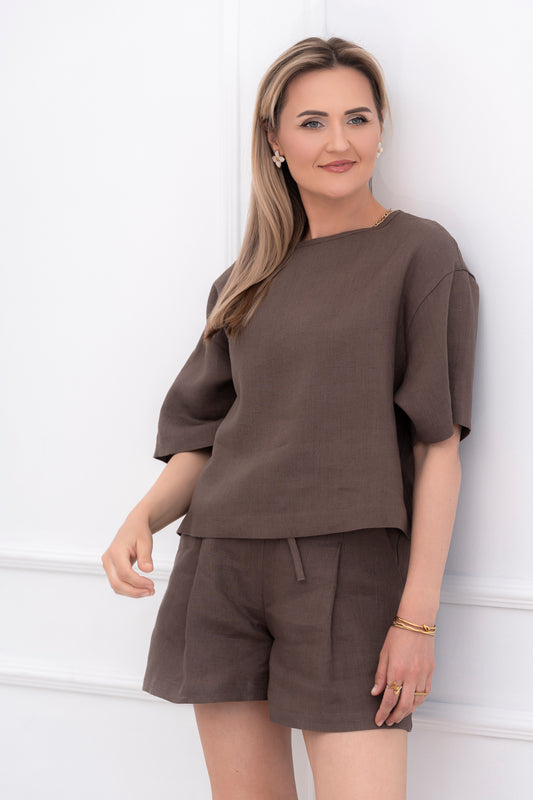 Lininė moteriška palaidinė, didinta, plati, trumpomis rankovėmis. Trumpa, laisvo modelio. Spalva - tamsiai ruda. Modelis taip pat vilki derančius lininius šortus.