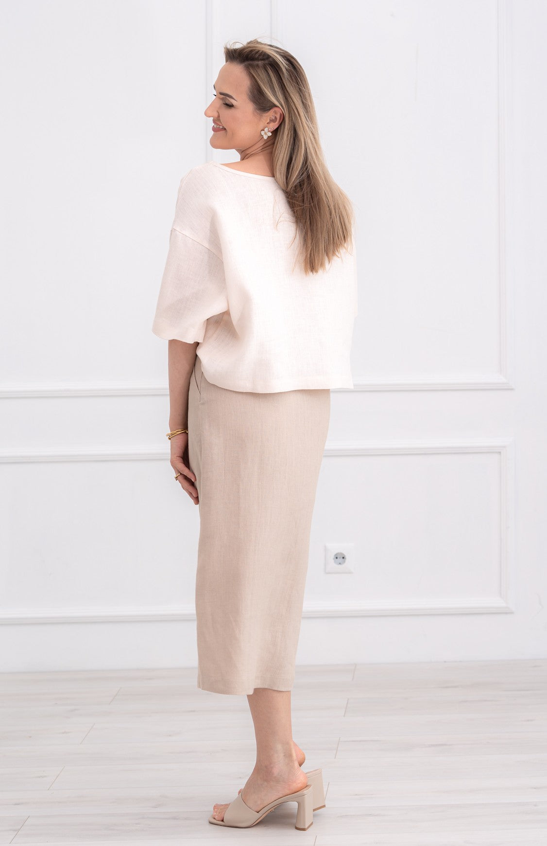 Lininė moteriška palaidinė, didinta, plati, trumpomis rankovėmis. Trumpa, laisvo modelio. Spalva - kreminė balta. Modelis taip pat vilki smėlio spalvos lininį sijoną