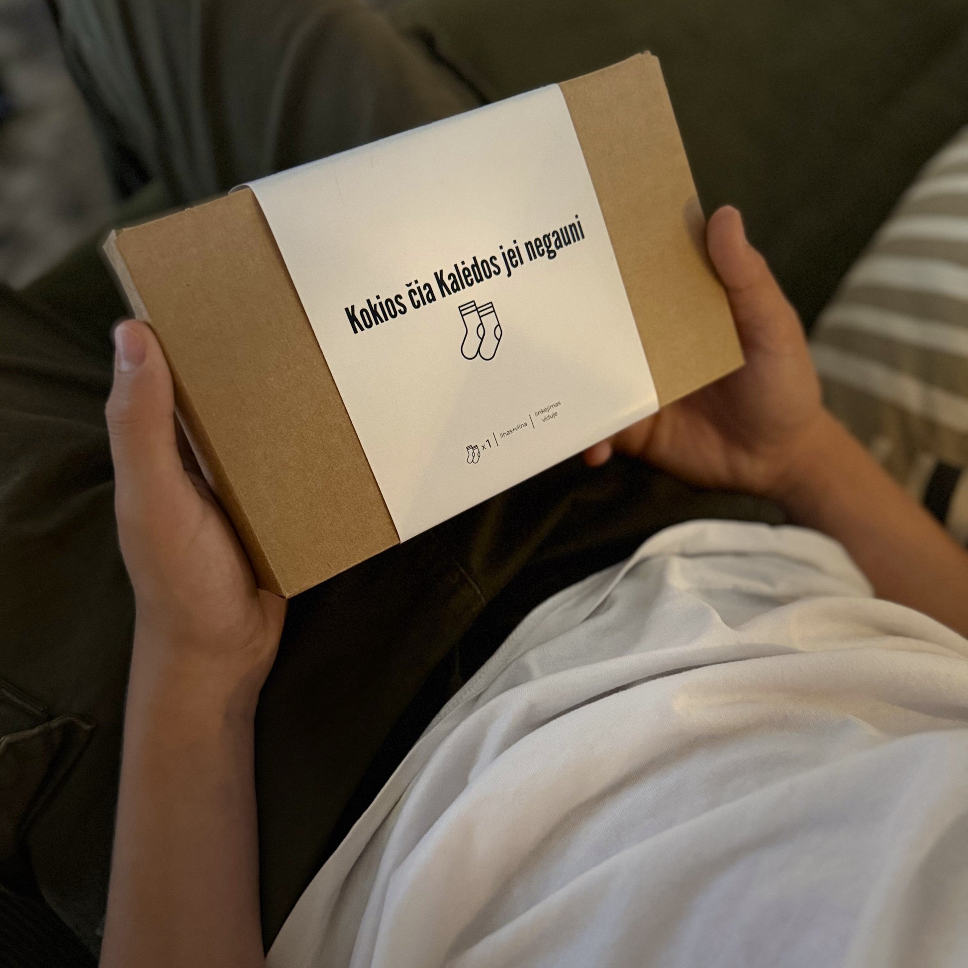 jaunuolis rankose laiko dėžutę, kurioje supakuotos kalėdinės baltos kojinės, lino-vilnonės kojinės. ant dėžutės užrašas Kokios čia Kalėdos jei negauni kojinių