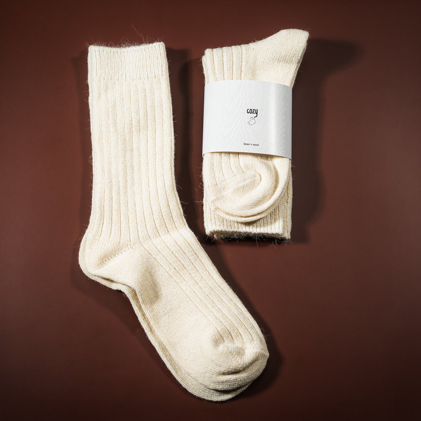 Lino-vilnos kojinė, kreminės baltos spalvos, su etikete cozy socks, ir šalia padėtos kojinės ištiestos be etiketės