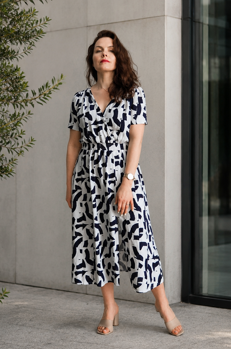 Woman wearing a flowing white and deep navy printed midi dress with short gathered sleeves, an elastic waist, and a side slit. Styled with dark heels against a minimal studio background.