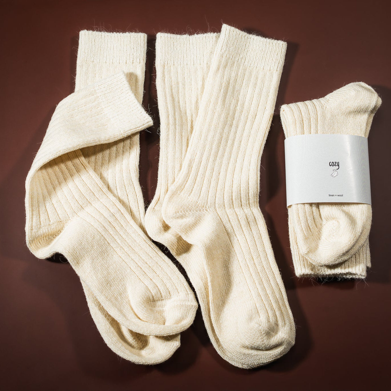 Lino-vilnos kojinė, kreminės baltos spalvos, su etikete cozy socks, ir šalia padėtos kojinės ištiestos be etiketės