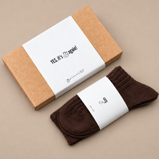 Tamsiai rudos lino ir vilnos kojinės šalia kraftinės dovanų dėžutės su užrašu „YES, it’s socks again!“ smėlio spalvos fone.